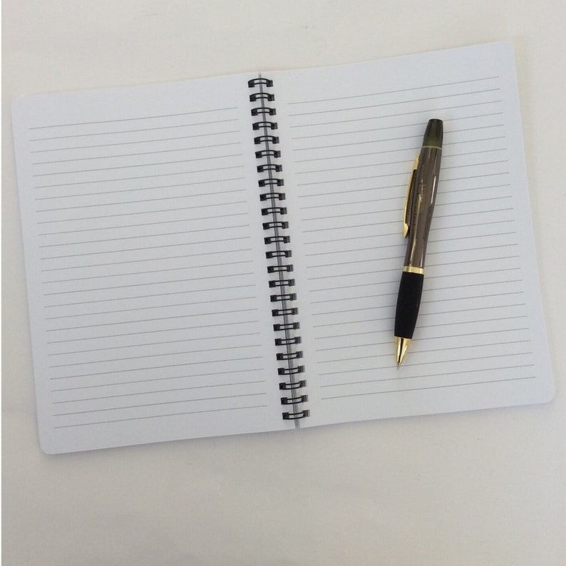 May include: An open, lined notebook with a black spiral binding and a pen. The pen is dark gray with gold accents and a black grip. The notebook's pages are white with horizontal lines.