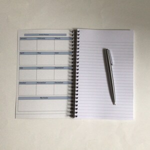 May include: Open spiral-bound planner with a yearly calendar layout and lined pages. A silver pen rests on the right page. The calendar includes months from January to December and a section for goals.