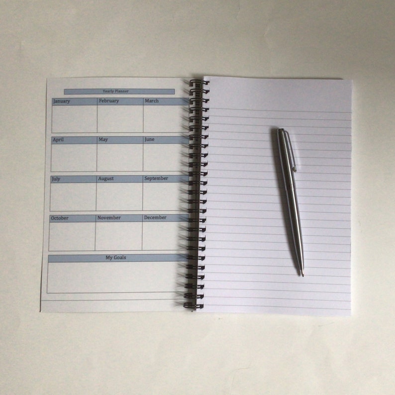 May include: Open spiral-bound planner with a yearly calendar layout and lined pages. A silver pen rests on the right page. The calendar includes months from January to December and a section for goals.