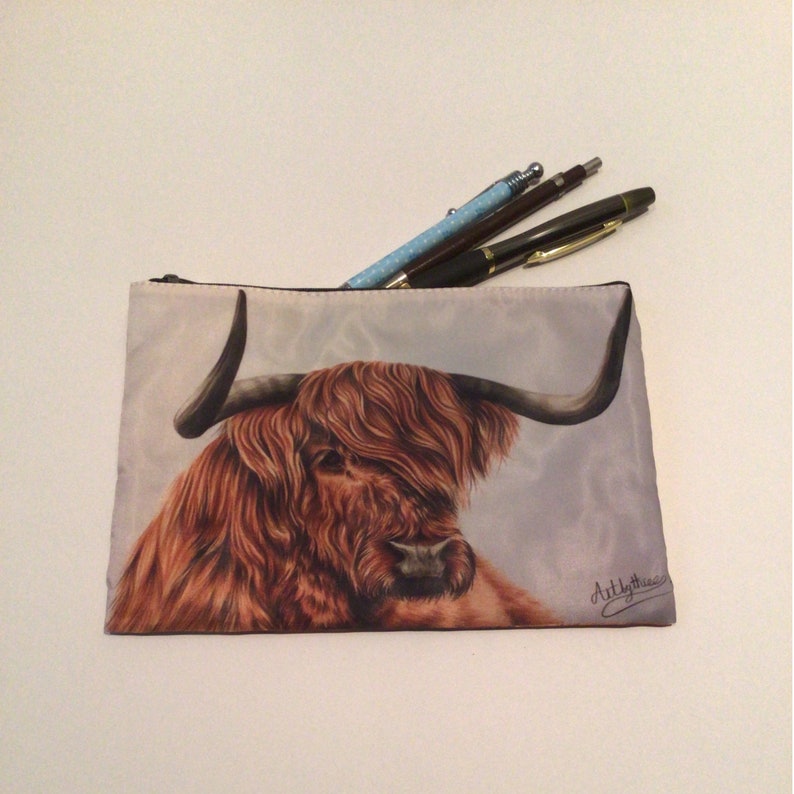 May include: A zippered pouch with a brown and white Highland cow illustration. The cow has long, shaggy hair and a large, curved horn. The pouch is made of a soft, fabric material and has a zipper closure. The pouch is perfect for storing small items like pens, pencils, and makeup.