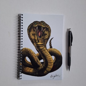May include: A spiral-bound notebook with a detailed illustration of a cobra, mouth agape, revealing fangs and tongue. The snake is depicted in brown and gold hues, with green eyes. A black pen is positioned next to the notebook.