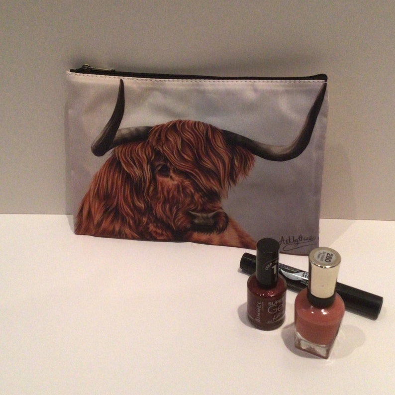 May include: A white zippered makeup bag with a brown and white Highland cow illustration. The bag is on a white surface with two bottles of nail polish and a black eyeliner.