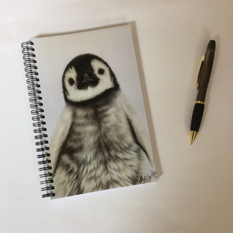 May include: A spiral-bound notebook featuring a detailed pencil drawing of a baby penguin. The penguin has black and white plumage and is centered on the page. A pen with a black grip and gold accents rests beside the notebook.