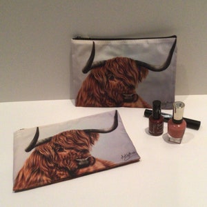 May include: Two zippered pouches with a brown and white Highland cow design. The pouches are made of a soft, fabric material.