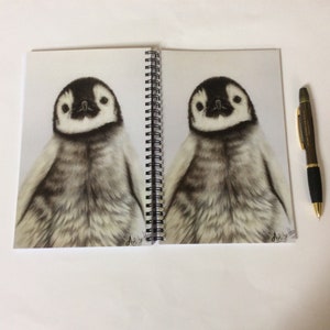 May include: A spiral-bound notebook featuring two detailed pencil drawings of a baby penguin. The penguin drawings are in black, white, and gray, with a pen resting on the right side. The notebook is open, displaying the artwork.