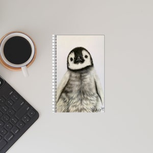 May include: A spiral-bound notebook featuring a detailed pencil drawing of a baby penguin. The penguin has a black head and back, with a white belly and gray shading. The notebook is on a white surface with a cup of coffee and a keyboard.