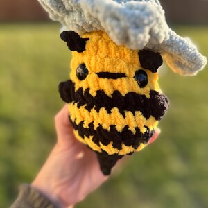 May include: A crocheted bee plush toy wearing a gray crocheted cloud hat. The bee is yellow and black with black eyes and a black mouth.
