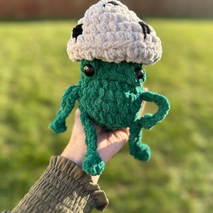 May include: A green crocheted plush toy with a white crocheted mushroom hat. The mushroom hat has black spots.