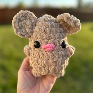 May include: A crocheted plush toy shaped like a mouse with large ears, brown fur, black eyes, and a pink nose.
