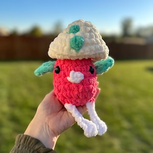 May include: A handmade crochet mushroom plushie. The plushie features a bright pink body, a white cap with teal accents, and teal arms. It has small black eyes and white legs. The plushie is held in a hand against a blurred green background.