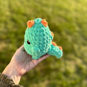 May include: A crocheted teal dinosaur toy with orange accents. The dinosaur has a small black eye and a long tail.