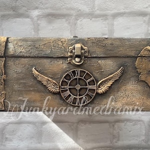 May include: A wooden box with a distressed gold finish and a metal clock face with wings. The box has a metal latch and is decorated with metal accents.