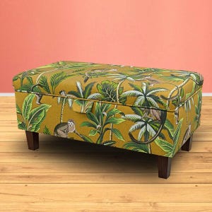 May include: A yellow storage ottoman with a tropical print featuring monkeys and palm trees. The ottoman has a hinged lid and wooden legs.