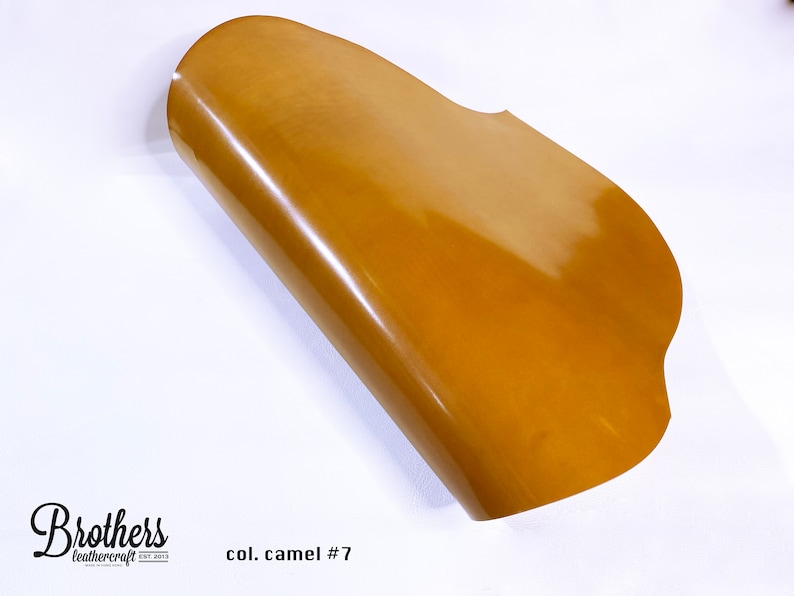 May include: A close-up of a piece of camel colored leather. The leather is folded in half and has a smooth, shiny finish. The text "col. camel #7" is visible in the lower right corner of the image.