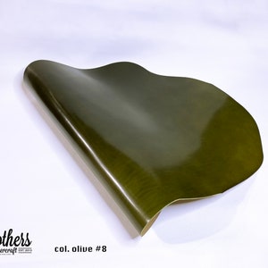 May include: A close-up of a piece of olive green leather. The leather is smooth and shiny, and it is folded in half. The text "col. olive #8" is printed on the white surface below the leather.