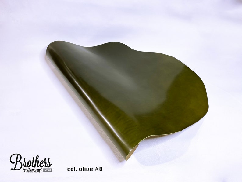 May include: A close-up of a piece of olive green leather. The leather is smooth and shiny, and it is folded in half. The text "col. olive #8" is printed on the white surface below the leather.