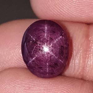Puede incluir: Una piedra preciosa de rubí estrella de color púrpura intenso y forma ovalada, que presenta un efecto de asterismo de seis rayos que irradia desde un punto central. La piedra tallada en cabujón tiene una superficie lisa y pulida.