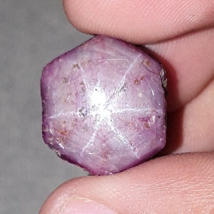 May include: A purplish-red gemstone with a six-pointed star pattern. The gem is held between fingers, highlighting its natural texture and colour variations. The star effect is prominent, reflecting light.
