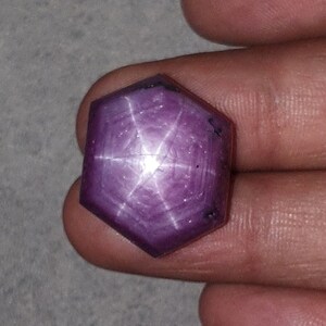 Pode incluir: Uma pedra preciosa de rubi estrela facetada com um padrão de estrela de seis pontas. A gema é de cor roxa escura com uma estrela branca brilhante no centro. A gema é segurada em uma mão.