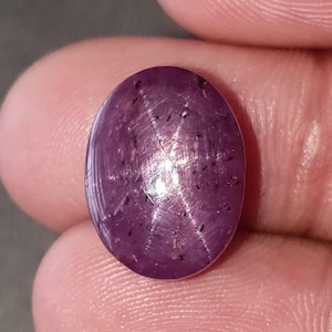 Può includere: Una gemma di rubino stellato di forma ovale, tagliata a cabochon, con una tonalità rosso violaceo intenso. La pietra mostra un distinto effetto a stella a sei raggi, noto come asterismo, centrato sulla superficie lucida, segnata da sottili inclusioni scure.