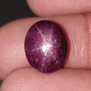 Può includere: Una gemma ovale bordeaux scuro con un motivo a forma di stella. La gemma è tenuta tra le dita, mettendo in mostra la sua superficie lucida e il gioco di luce sulle sue sfaccettature. Il colore della gemma è un rosso scuro intenso.