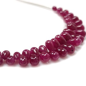 Könnte beinhalten: Eine Halskette mit einer Kette aus polierten, tiefroten Rubinperlen. Die Perlen sind tropfen- und rundförmig und auf einer dünnen, silberfarbenen Kette aufgereiht. Die Edelsteine sind in einem gebogenen Muster vor weißem Hintergrund angeordnet.