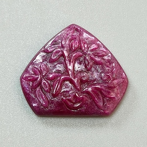 Könnte beinhalten: Ein geschnitzter, rubinroter Edelstein mit floralem Design. Der Edelstein hat die Form eines Schildes mit flacher Rückseite und einem geschnitzten Blumenmuster auf der Vorderseite. Die Schnitzerei zeigt Blumen und Blätter. Der Edelstein liegt vor einem hellgrauen Hintergrund.