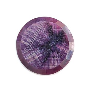 May include: A round, faceted purple gemstone featuring a distinct dark, cross-shaped inclusion at its center. The stone displays a translucent quality with visible internal striations and a polished surface, showcasing a deep violet hue.