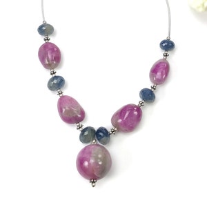 Puede incluir: Un collar con grandes piedras rosas ovaladas pulidas, intercaladas con piedras azul-gris más pequeñas y redondas. El collar está ensartado en una cadena plateada y tiene borlas blancas. El fondo es blanco con algunas flores.