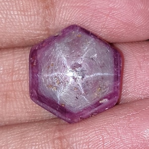 Op de afbeelding: Een gefacetteerde, zeshoekige edelsteen met een stervormig patroon. De edelsteen heeft een diepe roze-paarse kleur met een lichtere, bijna witte, stervormige insluitsel in het midden. De edelsteen wordt tussen vingers gehouden.