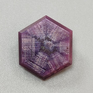 Könnte beinhalten: Ein sechseckiger, purpurroter Edelstein mit sternförmigem Muster. Der Edelstein hat eine polierte Oberfläche und eine komplexe innere Struktur. Der Stein liegt vor einem hellgrauen Hintergrund. Es handelt sich wahrscheinlich um einen Rubin.