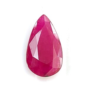 Puede incluir: Una piedra preciosa suelta en forma de pera de color rojo magenta intenso con una superficie facetada. La piedra presenta una parte superior puntiaguda y una base redondeada, mostrando múltiples facetas geométricas que reflejan la luz.