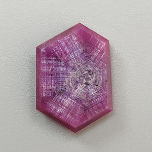 Può includere: Una gemma di rubino esagonale e sfaccettata, di colore rosa intenso. La gemma presenta un motivo a forma di stella sulla sua superficie lucida, che mostra una struttura interna unica. La gemma è posta su uno sfondo neutro.