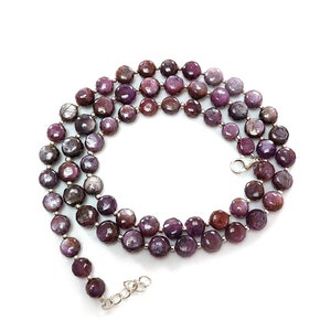 Puede incluir: Un collar con múltiples hilos de piedras preciosas facetadas de color púrpura intenso. Las cuentas están ensartadas con eslabones de metal plateado y un cierre. El collar se exhibe sobre una superficie blanca, con un arreglo floral en el fondo.