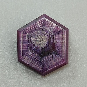May include: A faceted, hexagonal gemstone with a deep purple hue. The stone features intricate internal patterns and a polished surface, showcasing a blend of light and dark shades. The gem is set against a neutral background.