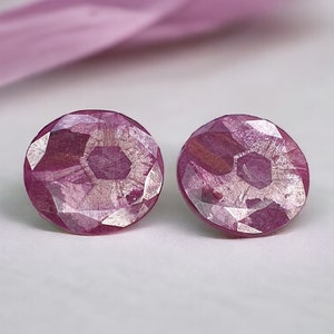 May include: A pair of round, faceted stud earrings in shades of pink and silver. The earrings have a geometric design and are set against a white background with a pink ribbon.