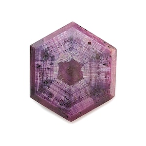 May include: A hexagonal, polished gemstone with a deep magenta hue. The stone features a central, star-like pattern and visible internal structures. The gem is set against a white background, highlighting its color and form.