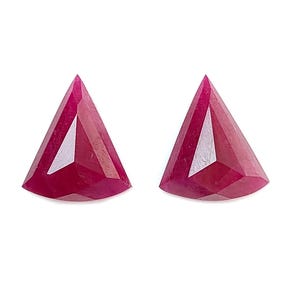May include: Two loose, vibrant red rubies cut into a triangular shape with multiple facets. The gemstones exhibit a deep, saturated crimson hue and a polished surface that reflects light across their geometric, pointed edges and flat bases.