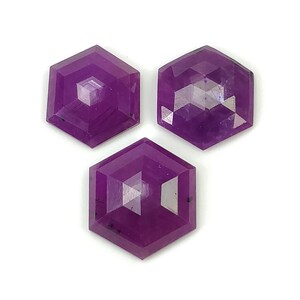 May include: Three loose, faceted gemstones with a distinct hexagonal shape and a deep purple colour. Each stone features a flat top surface with geometric facets that reflect light, showcasing a translucent quality and a uniform, saturated violet hue.