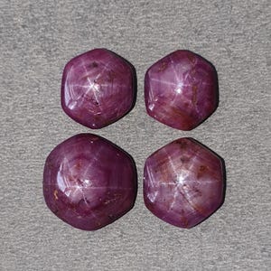 以下が含まれることがあります： 星の効果がある、ファセット加工された楕円形の濃い赤色の宝石が4つ。 宝石は灰色の表面に正方形に配置されています。 宝石は研磨された反射面を持っています。