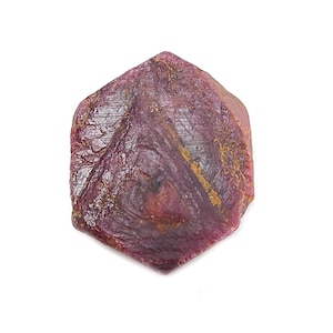 Puede incluir: Una gema de rubí en bruto, sin cortar, de color rojo púrpura intenso. La piedra natural tiene una superficie texturizada con líneas visibles y algunas inclusiones de color marrón dorado. El logotipo de Saksham Gems está en la esquina superior izquierda.