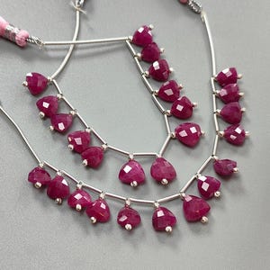 May include: Two strands of ruby gemstone beads with silver accents. Each strand features heart-shaped, faceted ruby beads. Pink tassels are attached to the top of the strands. The background is a solid grey.
