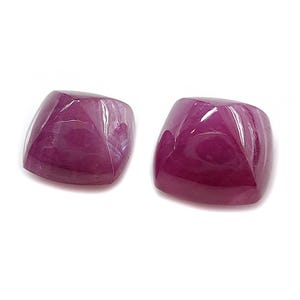 Könnte beinhalten: Zwei kissenförmig geschliffene, tiefrote Edelsteine mit polierter, reflektierender Oberfläche. Die Edelsteine sind quadratisch mit abgerundeten Ecken und vor weißem Hintergrund. Wahrscheinlich Rubine.
