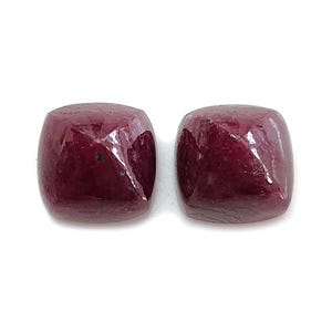 May include: Two polished, square-cut gemstones with a deep, rich burgundy color. The stones have rounded edges and a glossy surface, reflecting light. The gems are set against a white background.
