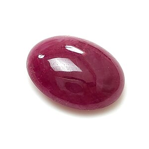 Pode incluir: Uma pedra preciosa de formato oval com corte cabochão e uma cor vermelho-magenta profunda e saturada. A superfície da pedra é lisa e polida, exibindo um brilho reflexivo que destaca o seu topo arredondado, convexo e opaco.