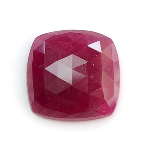 Pode incluir: Uma pedra preciosa de corte almofada com uma cor vermelho-magenta profunda e uma superfície facetada. A pedra apresenta uma forma quadrada simétrica com cantos arredondados e um acabamento polido que reflete a luz através das suas múltiplas facetas triangulares.