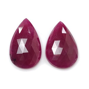 Könnte beinhalten: Zwei facettierte, tropfenförmige Rubine. Die Edelsteine haben eine tiefe, satte rote Farbe mit mehreren Facetten, die das Licht einfangen. Die Rubine werden vor einem weißen Hintergrund präsentiert, der ihre Farbe und Klarheit hervorhebt. Die Edelsteine sind wahrscheinlich für die Schmuckherstellung bestimmt.