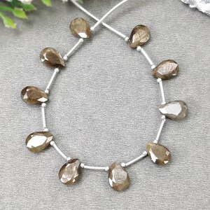 May include: A strand of ten faceted, pear-shaped brown gemstones arranged in a circular pattern on a silver-coloured wire. Each gemstone is separated by a small silver-coloured spacer bead, and the strand is finished with a silver-coloured tassel.