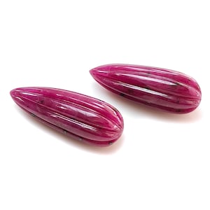 May include: Two polished, elongated ruby gemstones with a ribbed texture. The gemstones are a deep, rich red color, and are set against a white background. The shape is similar to a teardrop.