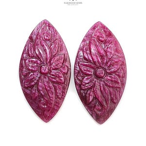 Op de afbeelding: Twee marquise-vormige, diep magenta gekleurde edelstenen met ingewikkelde, met de hand gesneden bloemmotieven op hun oppervlak. De stenen hebben een gepolijste afwerking met zichtbare natuurlijke insluitingen en texturen.